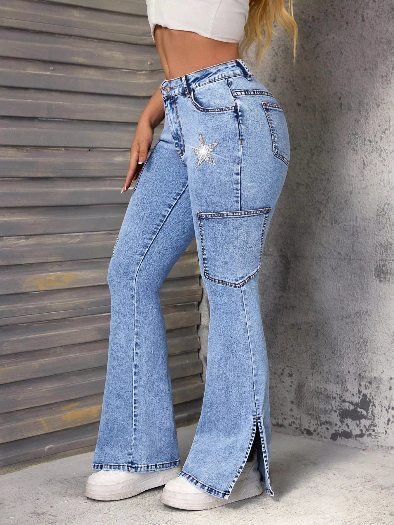 Lady wearing slit star flare leg blue denim jeans paired with a white tank top and white tennis shoes, with walls visible in the background.