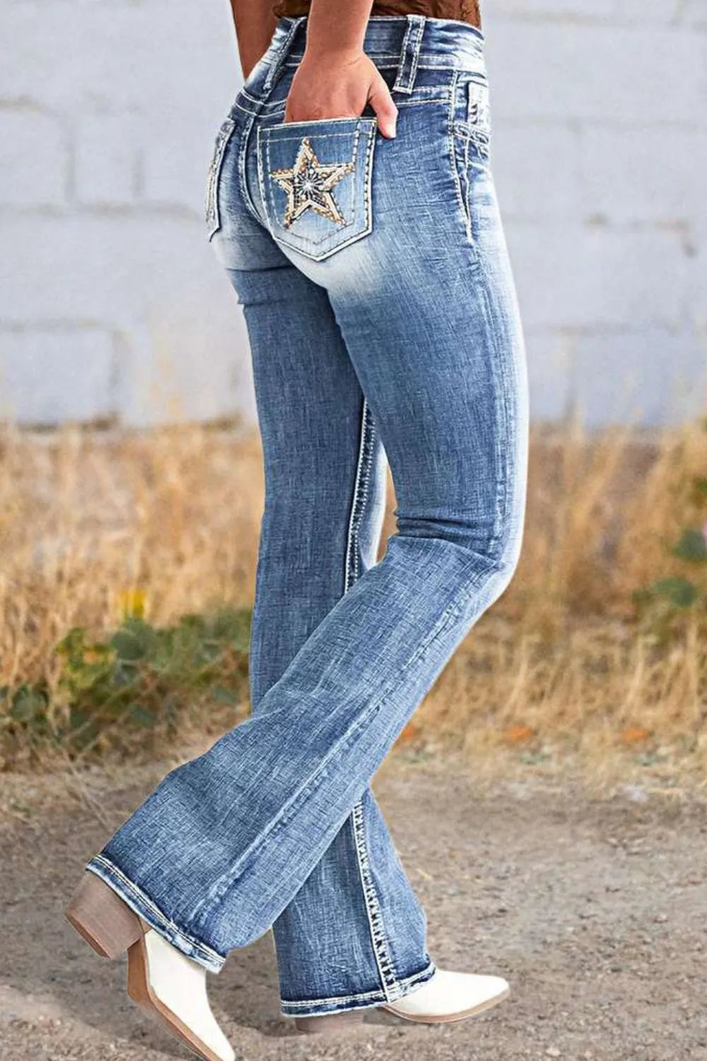 Woman wearing long boot cut acid wash blue jeans with star embroidered pockets, styled with sandals, with dried grass visible in the background.