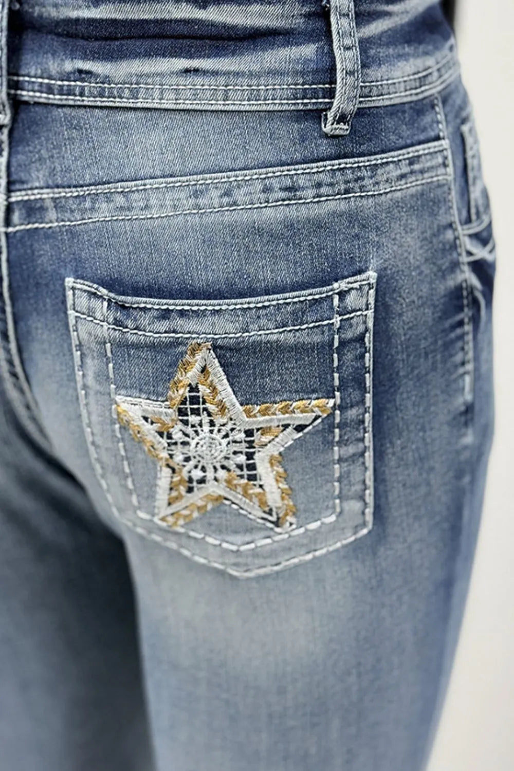 Woman wearing long boot cut acid wash blue jeans with star embroidered pockets, styled with sandals, with dried grass visible in the background.