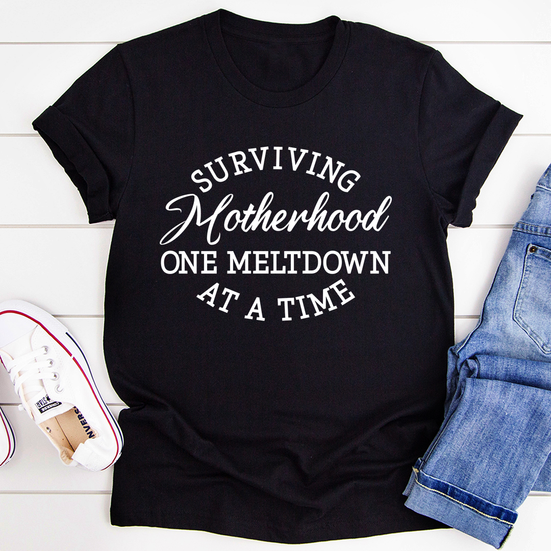 Black t-shirt with 'Surviving Motherhood One Meltdown At A Time' text on a white wooden surface with jeans and sneakers.