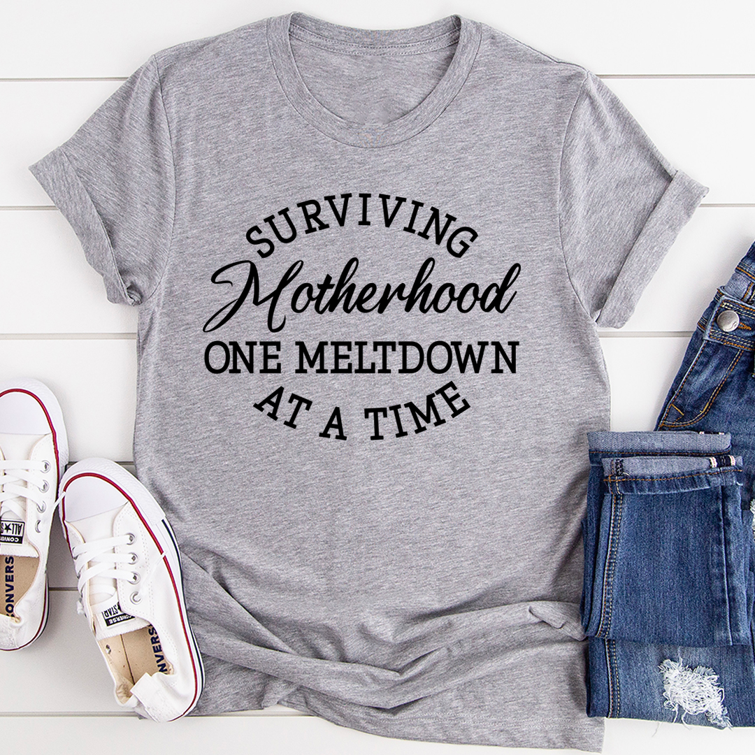 Gray t-shirt with 'Surviving Motherhood One Meltdown At A Time' text, paired with white sneakers and blue jeans on a light wooden surface.