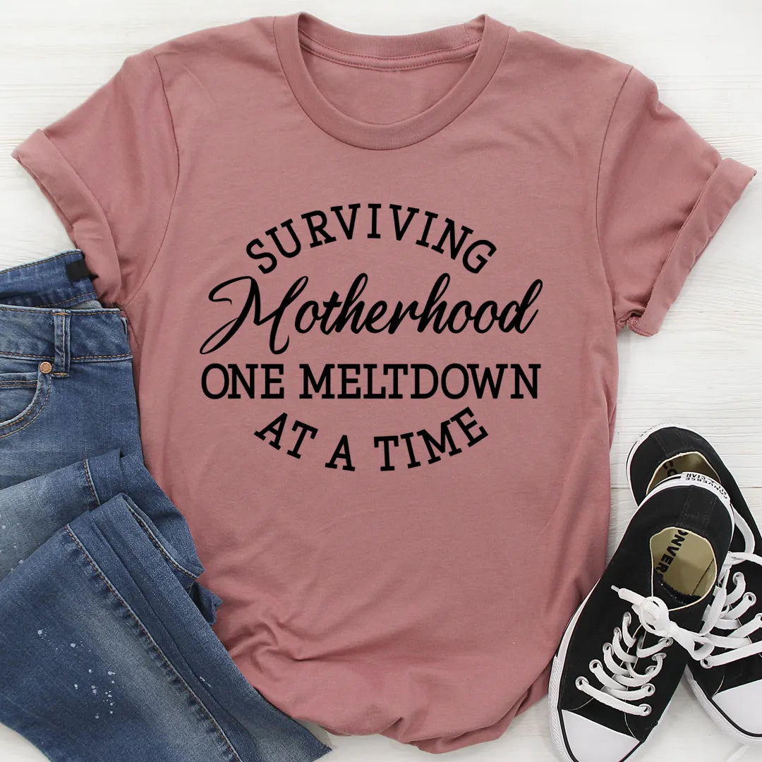 Pink t-shirt with 'Surviving Motherhood One Meltdown At A Time' text, paired with jeans and sneakers on a white background.