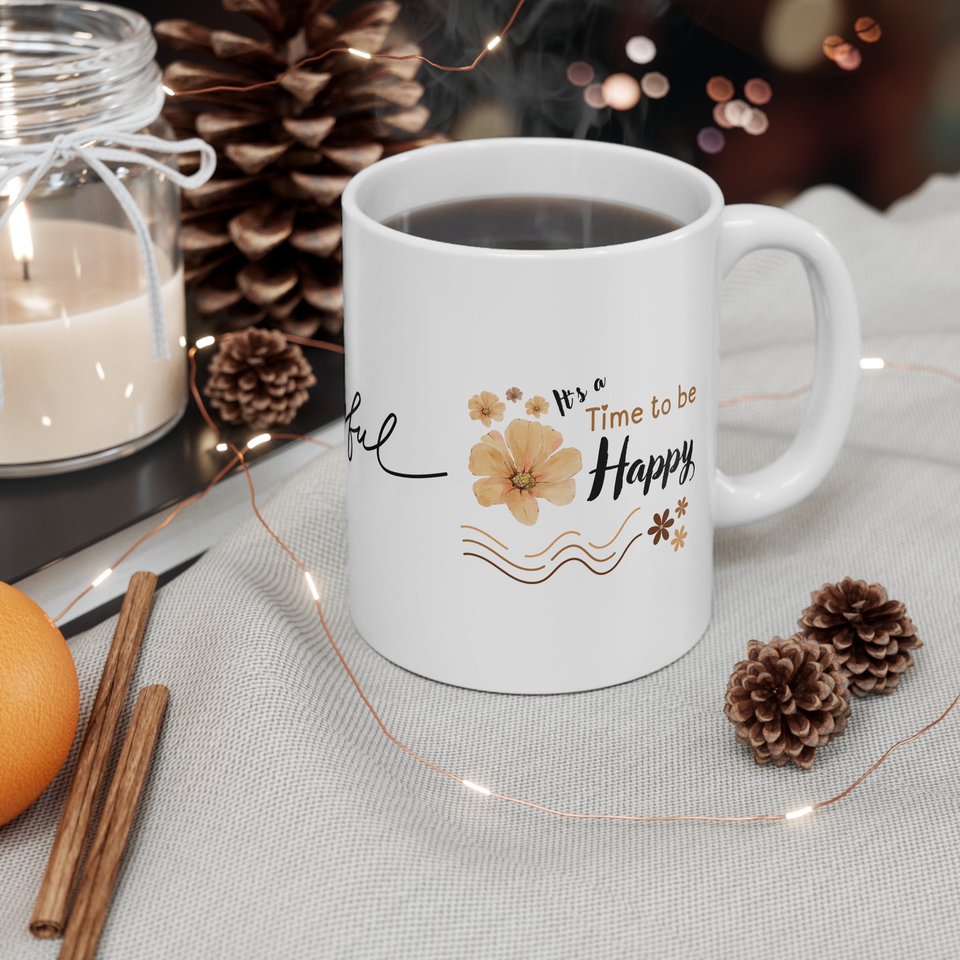 White mug with decorative text and flowers on a table with candles, oranges, and pinecones.