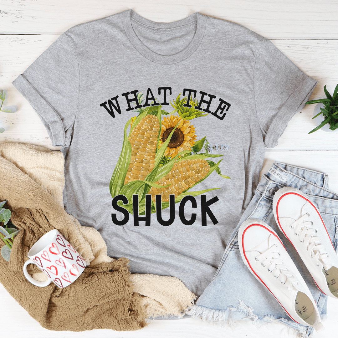 Gray t-shirt with corn and sunflower graphic and text 'WHAT THE SHUCK' on a white surface with casual items.