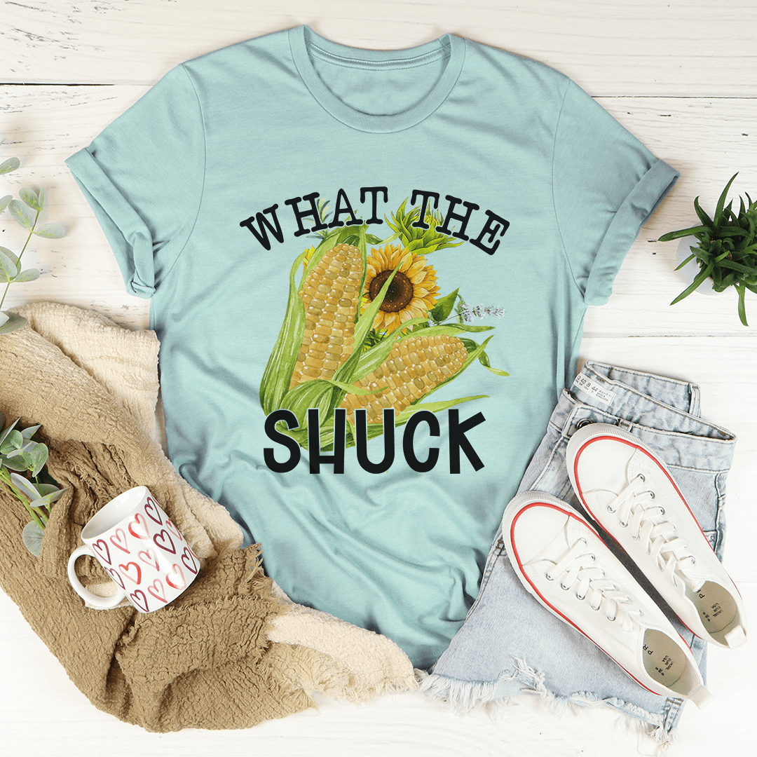 Light blue t-shirt with corn and sunflower graphic and text 'WHAT THE SHUCK' on a wooden surface with casual items.