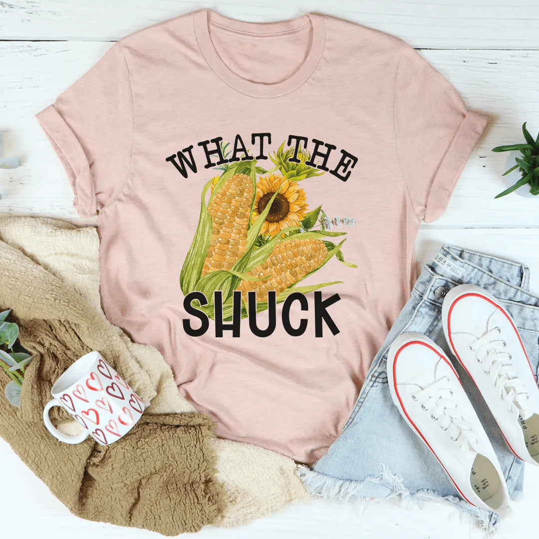 Pink t-shirt with corn and sunflower graphic and text 'WHAT THE SHUCK' on a white surface with casual items.