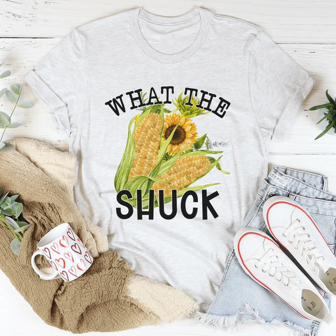 White t-shirt with corn and sunflower graphic and text 'WHAT THE SHUCK' on a white surface with accessories.