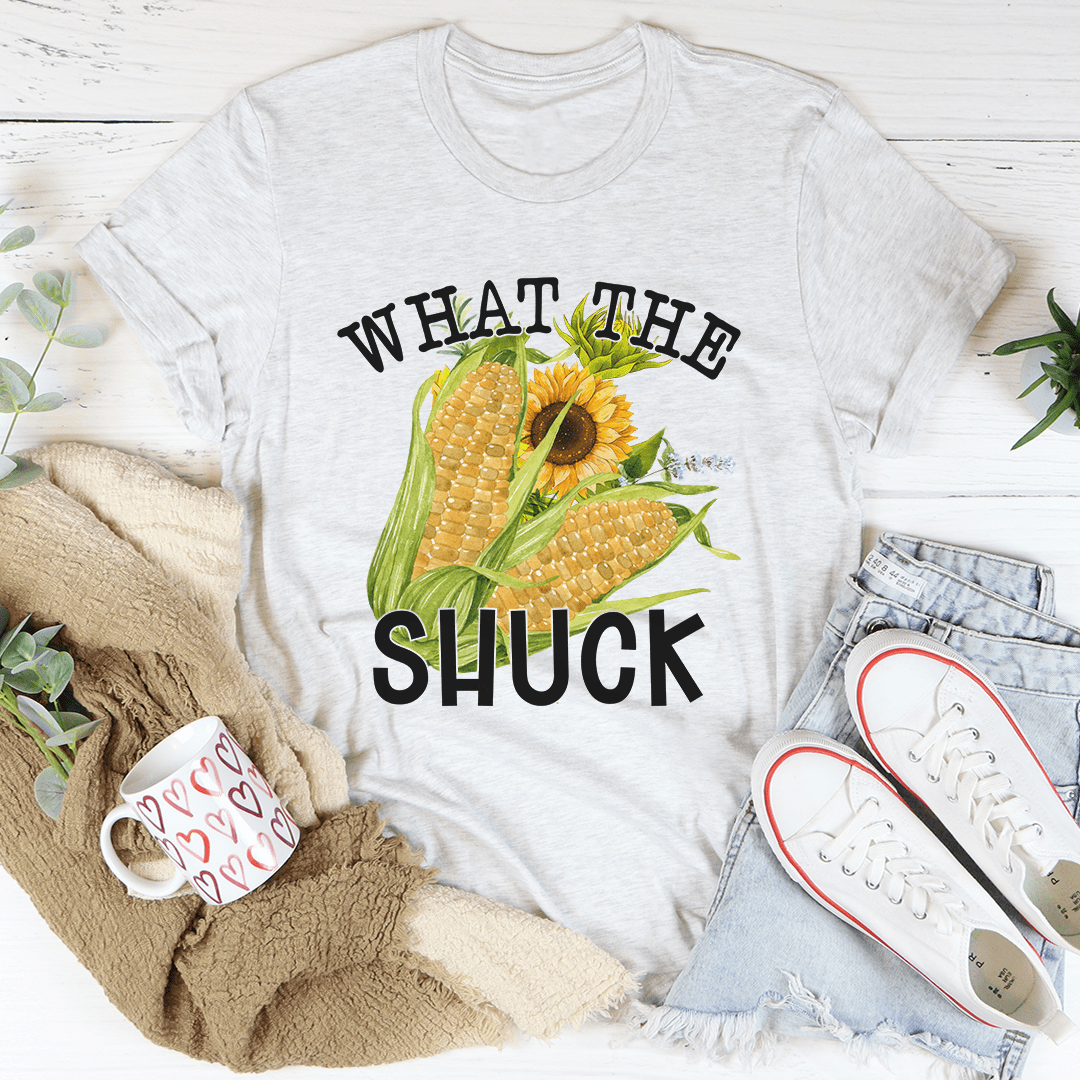 White t-shirt with corn and sunflower graphic and text 'WHAT THE SHUCK' on a white surface with casual items.