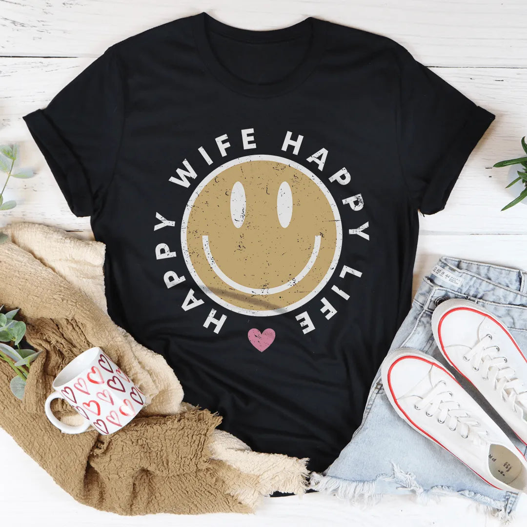 Black t-shirt with a smiley face and text on a wooden surface with casual items.