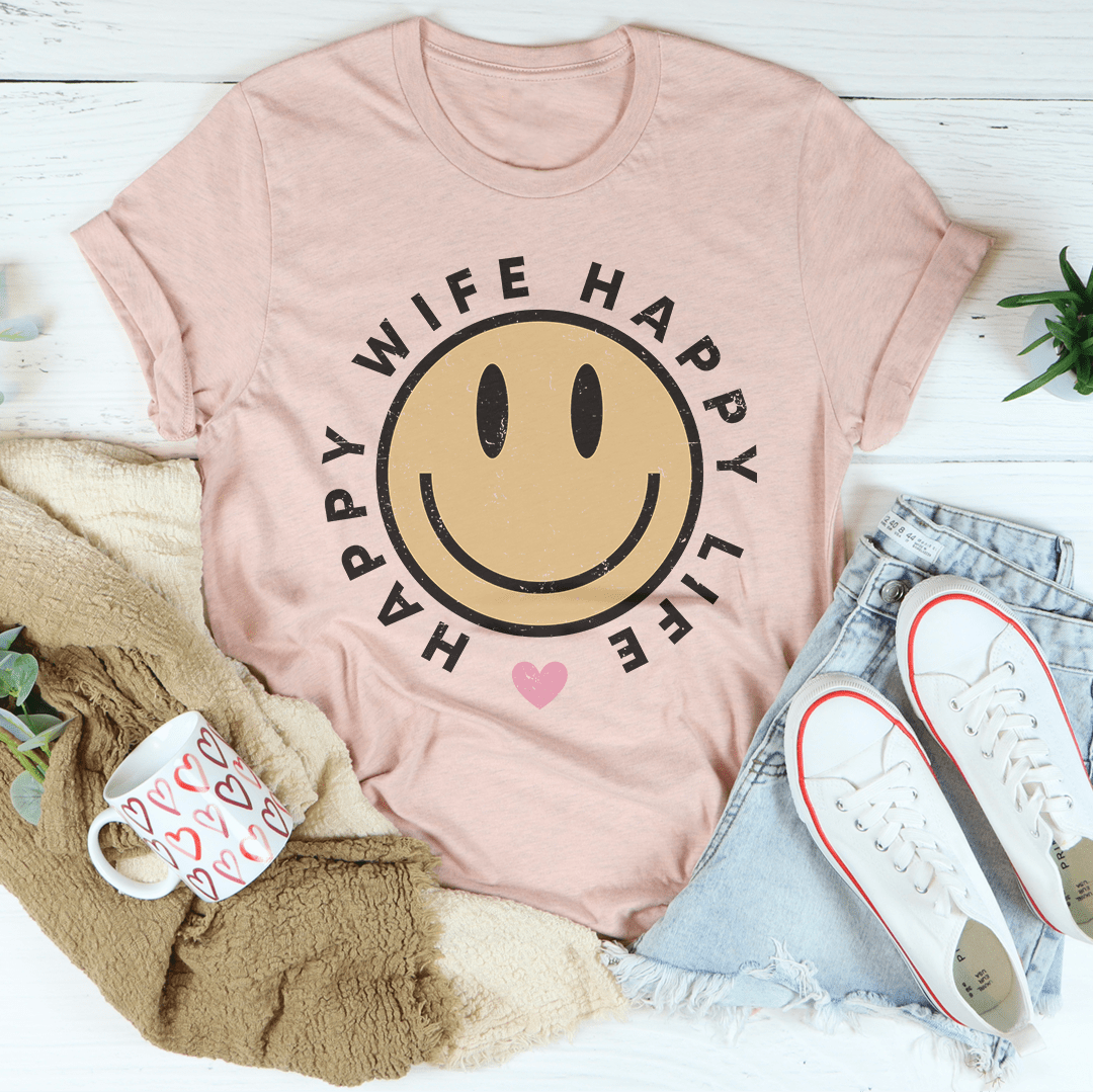 Pink t-shirt with smiley face design and text, placed on a surface with a mug, blanket, and shoes.