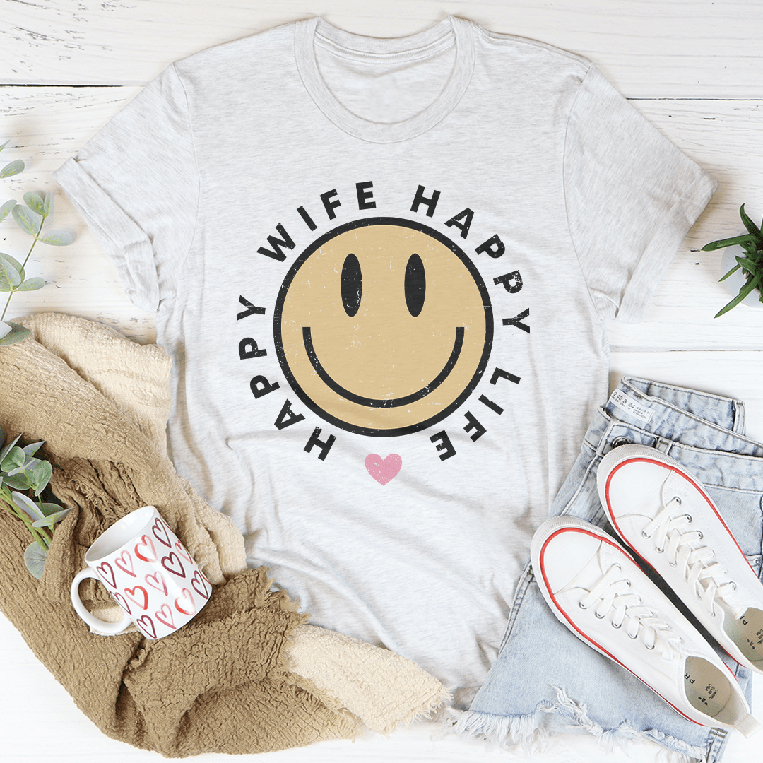 White t-shirt with smiley face design and text, placed on a white surface with casual items around.